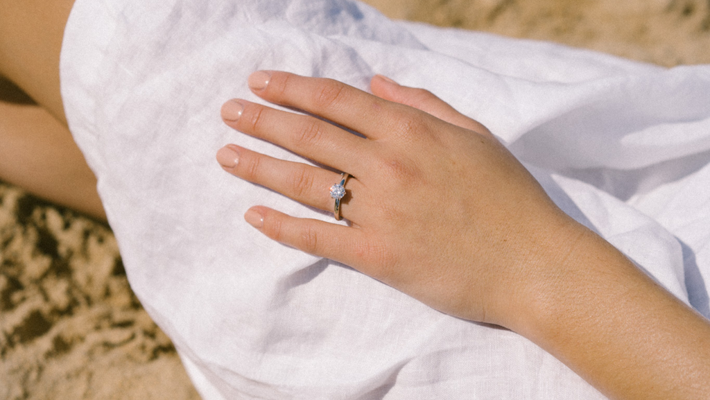 Everspark Moissanite engagement ring on the hand of a young lady who is sitting on the beach on the sand in a white dress